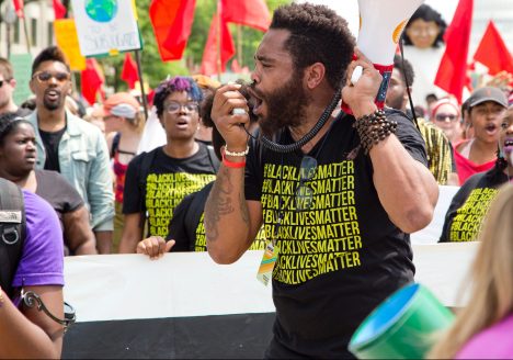 Black Lives Matter activist in black and yellow shirt with a bullhorn, surrounded by protesters with red flags and packards.